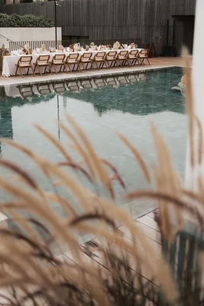 Reception tablescape on an outdoor reception table with dried florals, arranged poolside beside a modern wood wall and wooden deck.