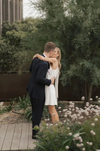 Wedding kiss portrait capturing a couple’s embrace, bride in a bridal mini dress, on a wood deck amid garden greenery and bright wildflowers.