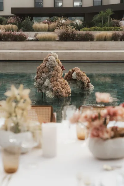Floating floral installation of hydrangeas drifts above a reflecting pool, with soft candles nearby, in a landscaped garden beside a modern facade.