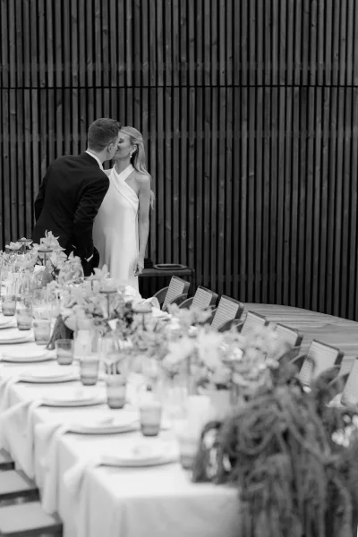 Wedding kiss portrait as the bride and groom kiss beside elegant floral centerpieces, set against a wood slat wall on a wooden deck.