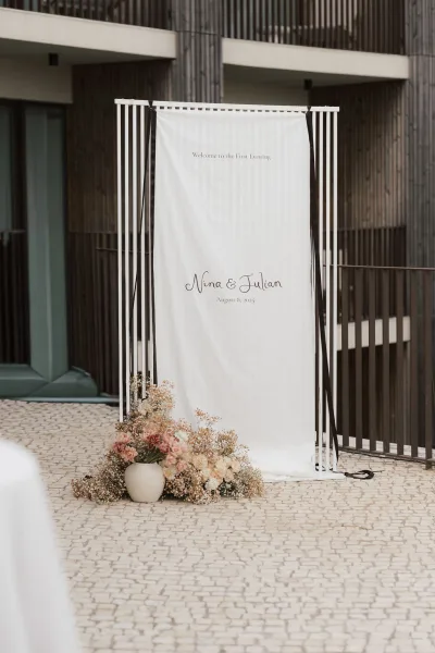 Wedding welcome sign as modern wedding signage on a white fabric banner, set in a paved courtyard by a modern building facade.