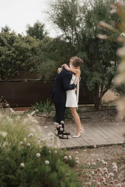 Couple portrait as the bride and groom hug in a garden, the bride in a bridal mini dress, framed by shrubs beside a wooden deck.