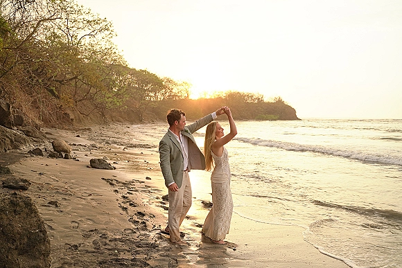Couple portrait of bride twirling in a lace wedding dress as groom leads, with boutonniere, at sunset by ocean waves and rocks