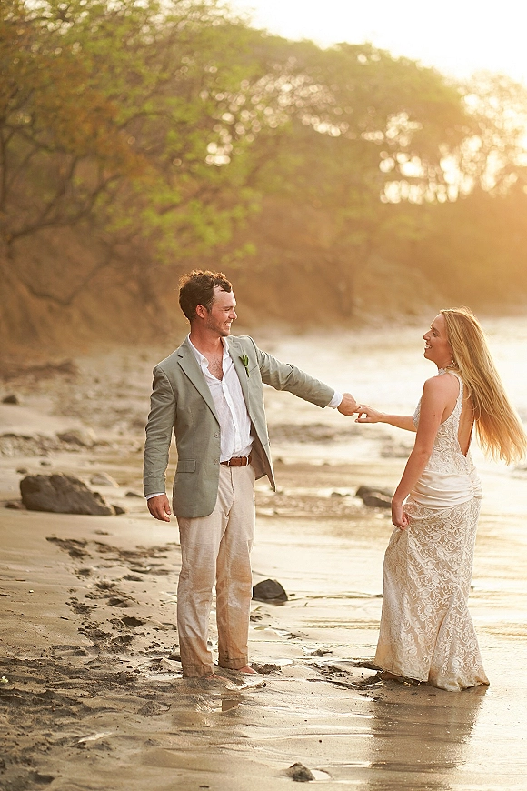 Couple portrait of bride and groom holding hands on a beach wedding couple walk along wet sand by rocks in golden sunset light