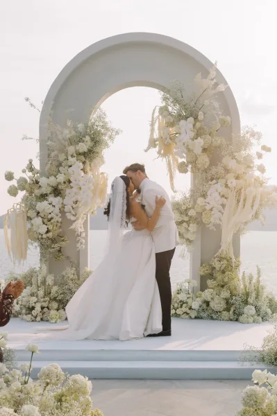 Wedding kiss beneath a modern white floral arch with orchids and baby's breath, bride’s veil flowing against ocean and sky backdrop
