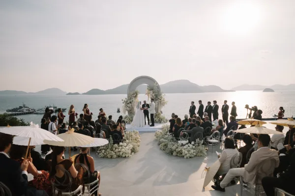 Wedding ceremony with bride and groom at a circular white floral arch on an oceanfront terrace at sunset, guests holding parasols