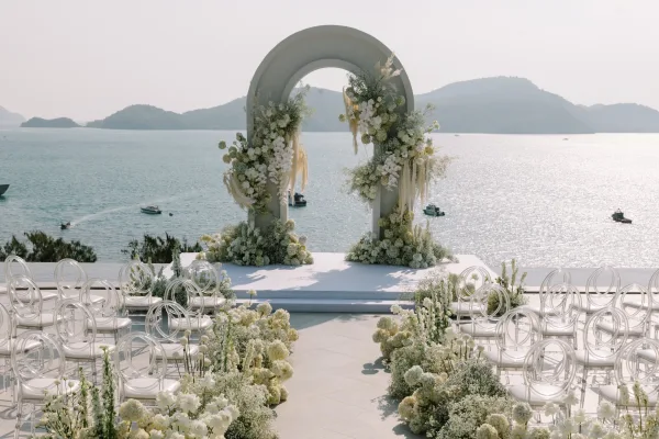 Ceremony setup with an oceanfront wedding ceremony floral arch backdrop, white and green flowers on a stage with acrylic chairs and aisle blooms, ocean and mountains behind