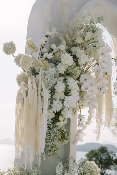 Wedding ceremony arch with a white floral arch of hydrangeas, roses, and orchids, set outdoors with mountains and water behind