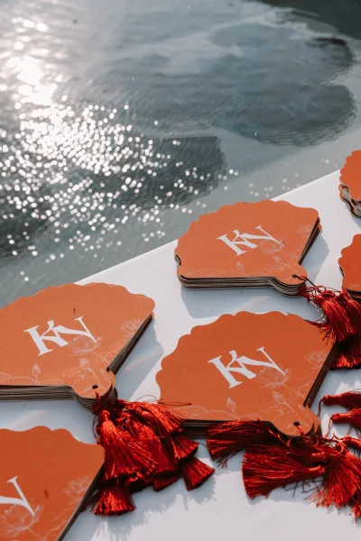 Wedding favor tags with wedding monogram tags and red tassels on scalloped cards tied with string, set on white linen near sunlit water reflections