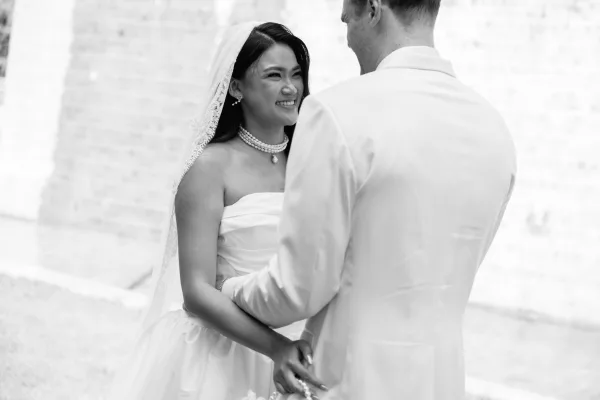 Ceremony moment as bride and groom holding hands, smiling during vows, with bridal veil and pearl necklace against a stone wall