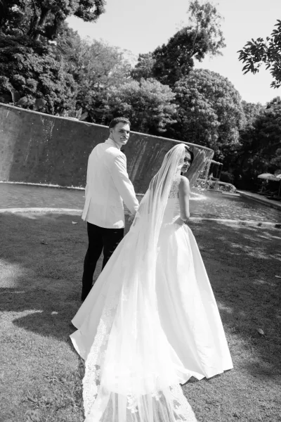 Couple portrait of bride and groom holding hands, walking away by a garden reflecting pool and waterfall, her cathedral veil trailing