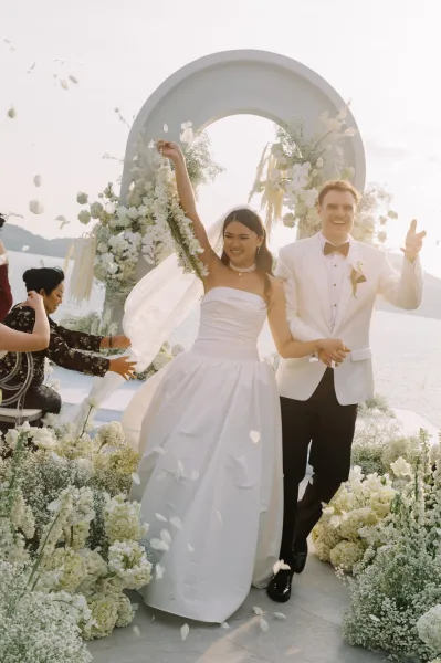 Recessional moment as bride and groom walk hand in hand through a rose petal toss beneath a white floral arch by a sunlit lake and mountains