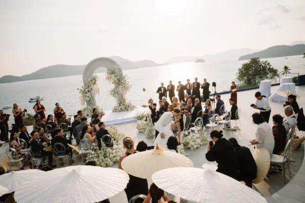 Ceremony kiss moment as the couple dips under a white floral arch, guests cheering beside clear chairs on a seaside terrace at sunset