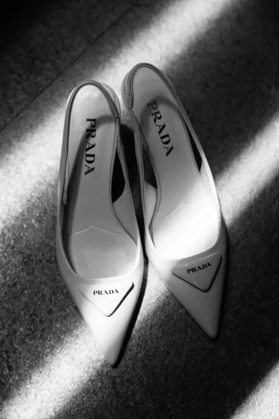 Bridal shoes flatlay of white pointed-toe heels with logo plaque on carpet floor, framed by soft window light shadows