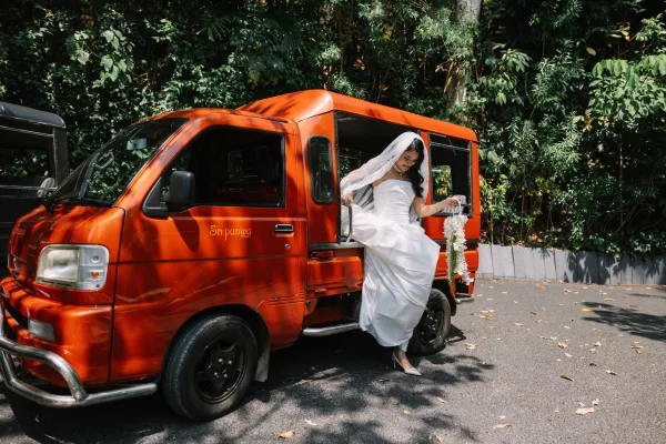 Bridal portrait of a bride in veil stepping from a red tuk tuk, holding a cascading orchid bouquet amid tropical greenery on pavement