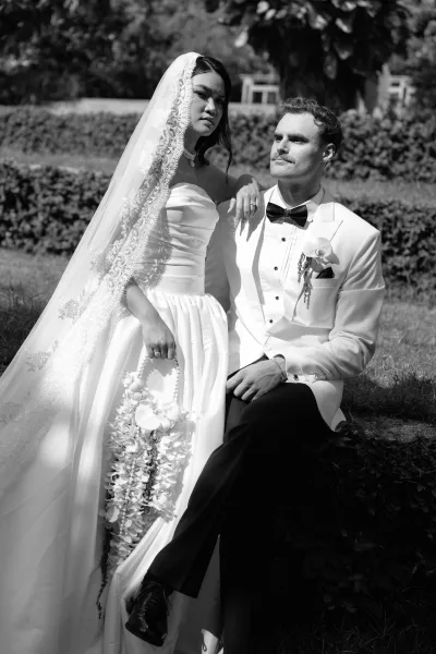 Couple portrait in a black and white wedding portrait style, bride standing with lace veil and orchid bouquet beside groom seated on lawn in sunlight