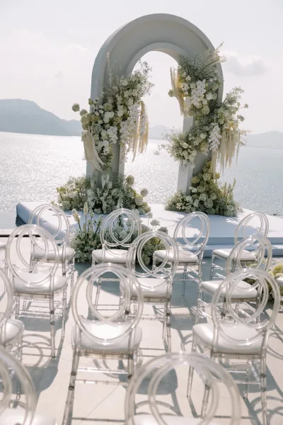 Ceremony setup for an outdoor wedding ceremony with a modern arched backdrop, white florals, and clear chairs overlooking a mountain lake
