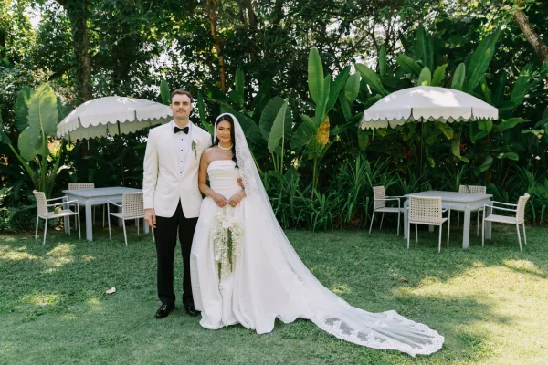 Couple portrait of bride in strapless dress with lace veil and cascading white bouquet beside groom in white tuxedo on tropical patio