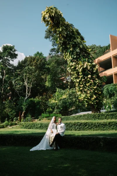 Couple portrait of bride and groom sitting on a garden lawn, bride in strapless gown with veil and cascading bouquet beside groom in tuxedo