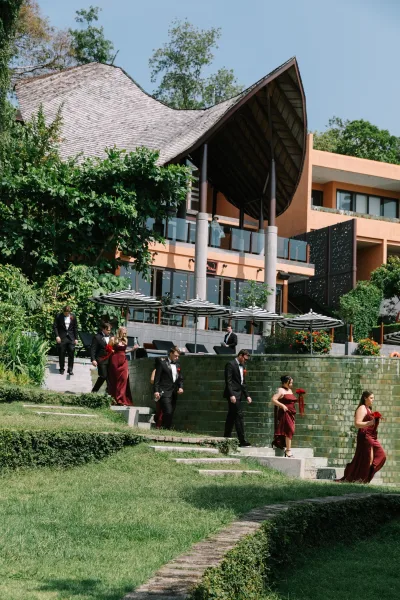 Wedding party processional with bridesmaids in burgundy dresses and tuxedoed groomsmen walking down stone steps at a modern resort pavilion with striped umbrellas