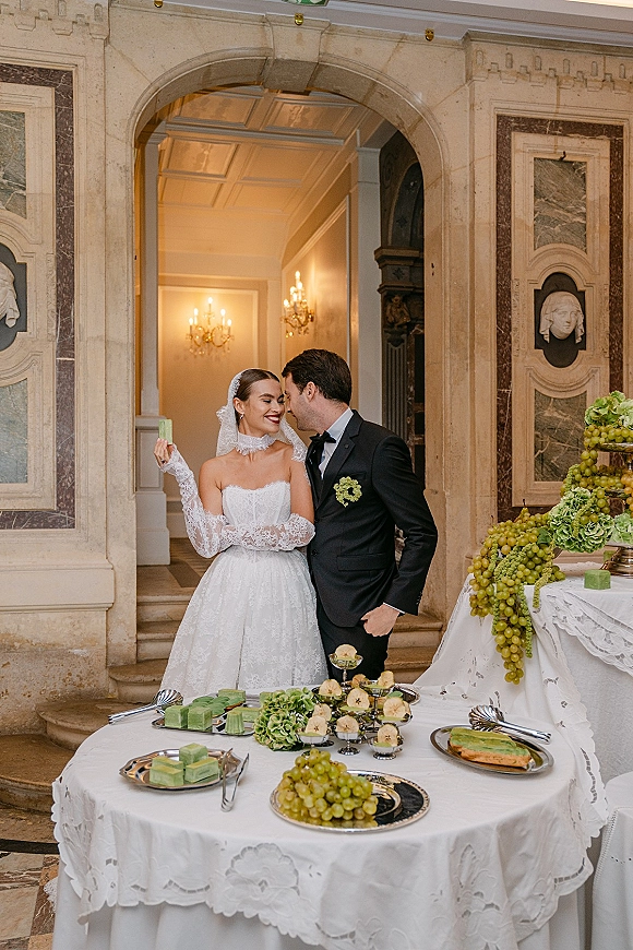 Couple portrait of bride and groom standing together under a stone archway, bride in strapless lace dress and veil by a green dessert table