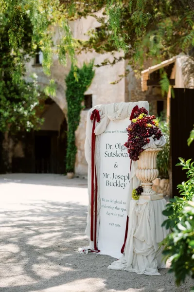 Wedding welcome sign with fabric draped signage set beside a stone archway along a gravel path amid garden greenery and trees.