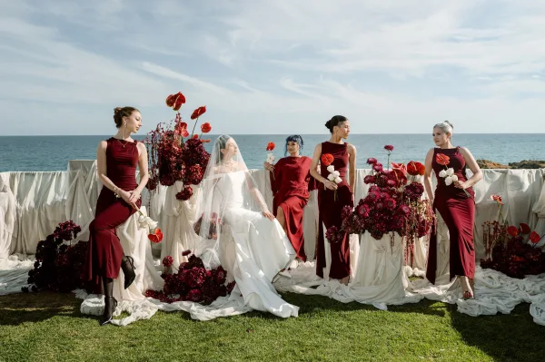 Bridal party portrait of bride with bridesmaids in burgundy gowns, holding red anthuriums, posed on a lawn by the ocean.