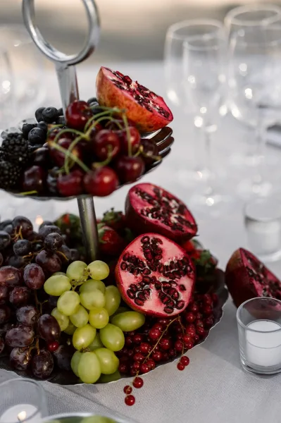 Fruit centerpiece on a tiered fruit stand filled with pomegranates, cherries, grapes, and red currants, accented by glowing votive candles.