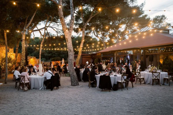 Outdoor reception decor for a string light wedding reception in a gravel courtyard beside a pavilion and pine trees at dusk.