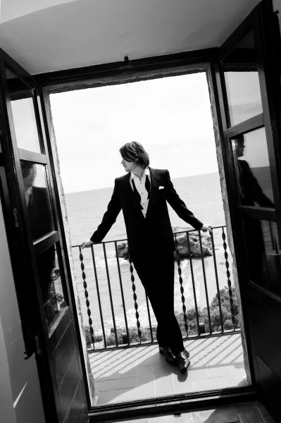 Groom portrait in black and white, wearing a tuxedo, standing on a balcony framed by open doors with the ocean coastline beyond.