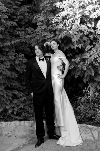 Couple portrait in black and white, bride in an off-the-shoulder gown with a pearl necklace beside the groom, framed by ivy and stone.