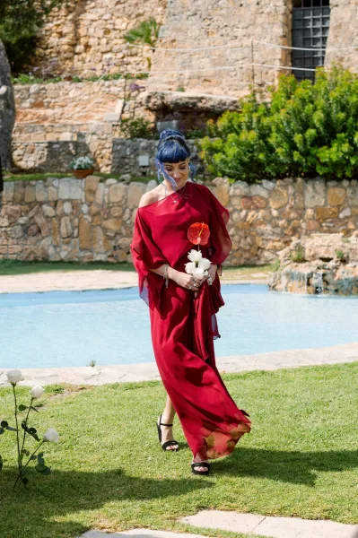 Ceremony processional during an outdoor ceremony moment, a figure in a red gown walks by a pool, stone wall, and greenery on the lawn.
