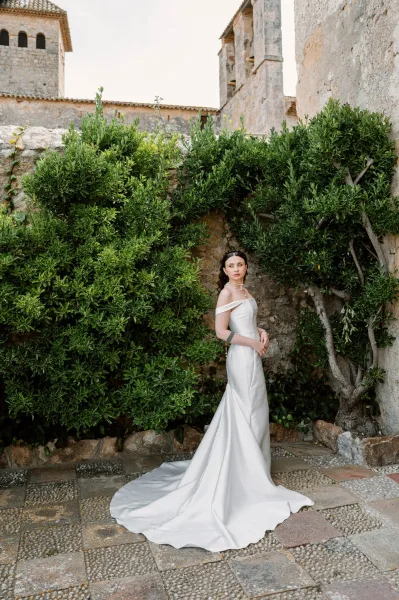 Bridal portrait of a bride in an off the shoulder wedding dress with a pearl necklace, set against a rustic stone wall and courtyard greenery.