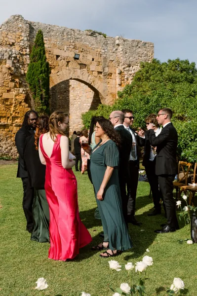 Wedding cocktail hour with guests mingling, drinks in hand, by a stone wall and archway bordering hedges under an open sky.