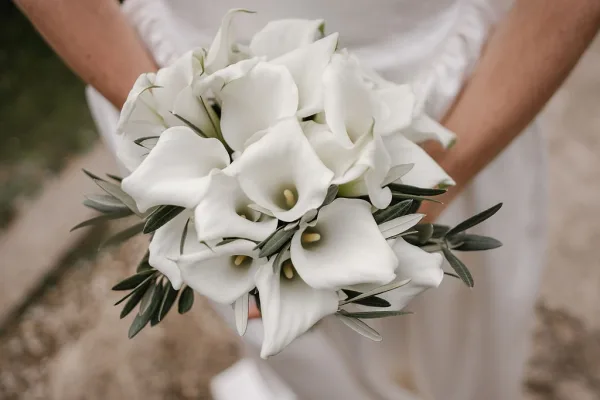 Bridal bouquet of white calla lilies with greenery accents held against a bridal dress, stems visible with softly blurred outdoor ground behind
