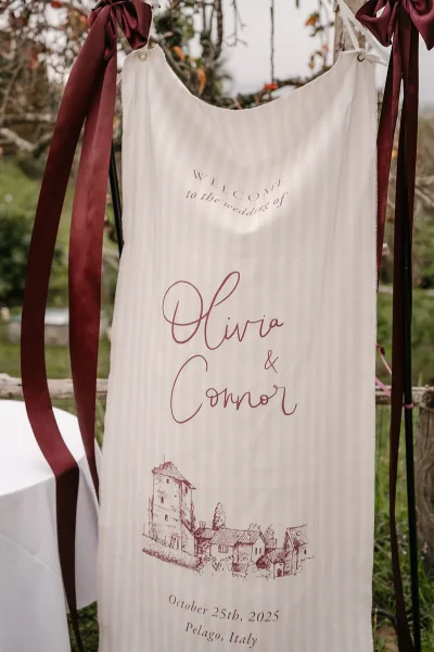 Wedding welcome sign in calligraphy on hanging fabric with burgundy ribbons, displayed on a black metal stand along a stone path outdoors