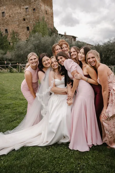 Bridesmaid group photo of the bride with bridesmaids in mix-and-match dresses, hugging on a lawn by a stone building under clouds