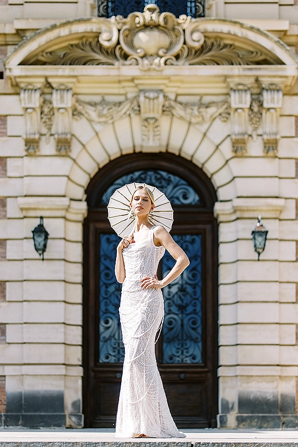 Bridal portrait of a bride in a beaded wedding dress holding a parasol on stone steps before an ornate arched doorway with iron door