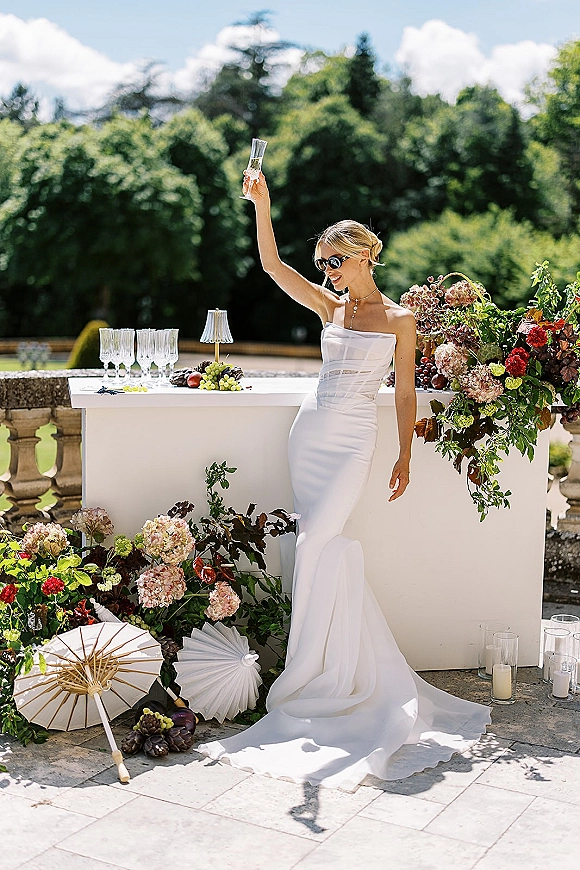 Bride portrait in a strapless wedding dress wearing sunglasses, holding a champagne flute at an outdoor terrace bar with florals and fruit