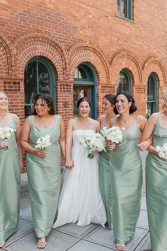 Bride with bridesmaids walking and laughing, carrying white bouquets in sage green dresses outside a brick building with arched windows