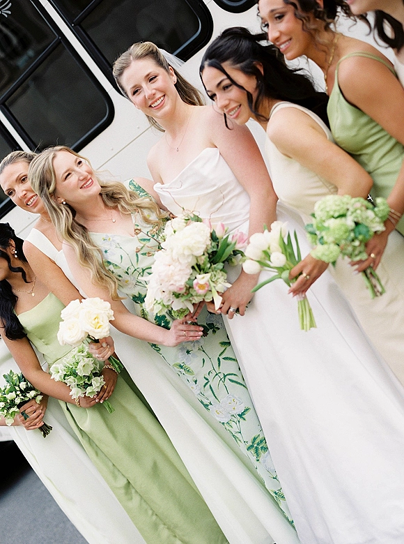 Bride and bridesmaids pose in a bridal party portrait, smiling with veils and white-green bouquets beside a white vehicle