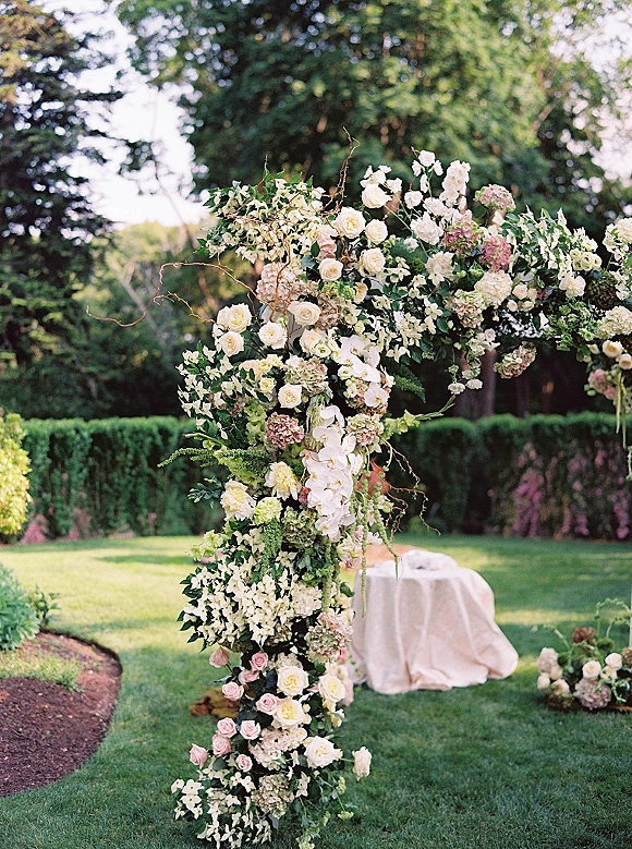 Wedding ceremony arch with roses and hydrangeas, asymmetrical greenery and orchids framing a garden lawn with hedges and trees behind