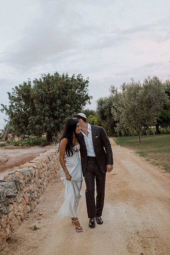 Couple portrait of bride and groom walking away as he kisses her forehead on a rustic dirt road beside a stone wall under cloudy skies