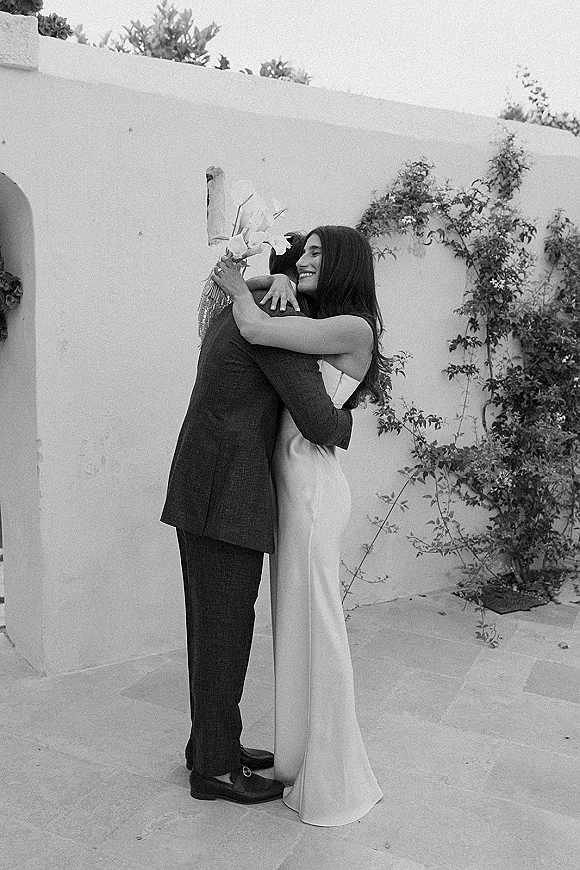Couple portrait of bride and groom in a wedding hug photo, her satin gown and white bouquet against a vine-covered stucco courtyard wall