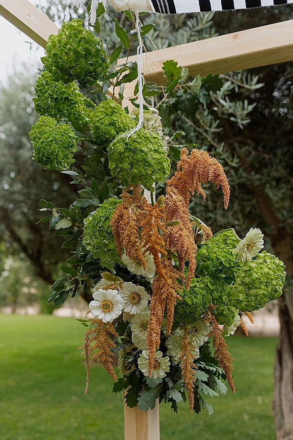Wedding arch flowers with an asymmetrical floral arrangement of green hydrangea, white blooms, greenery, and dried amaranth on a wooden arch in a garden lawn