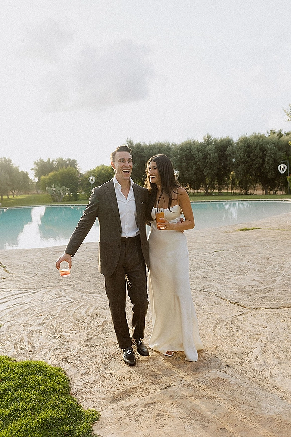 Couple portrait of newlyweds laughing with cocktail glasses on a stone patio by the pool, bride in strapless dress and groom in dark suit