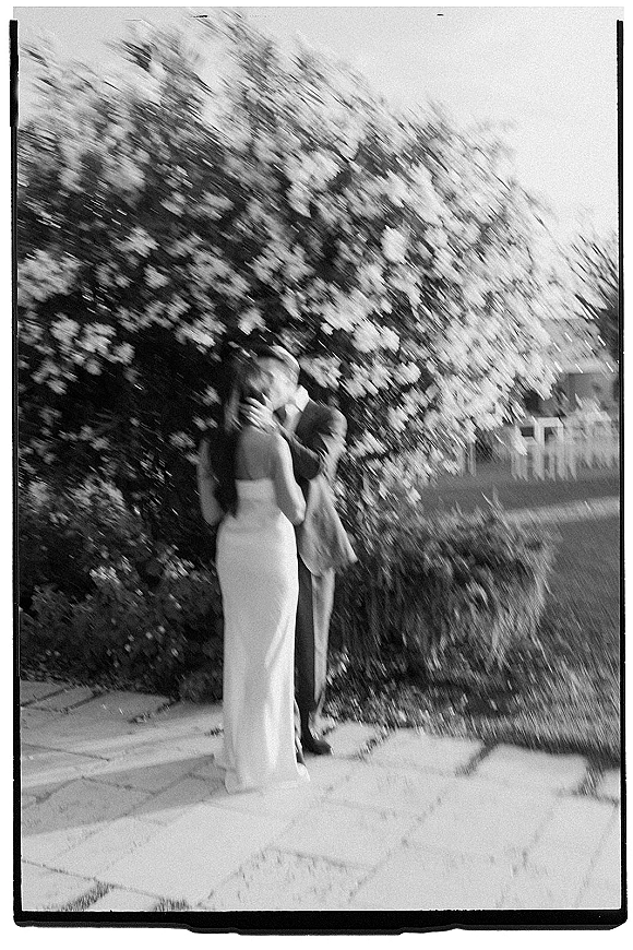 Wedding kiss portrait of bride and groom kissing on a stone patio, her strapless gown and his boutonniere framed by flowering bushes