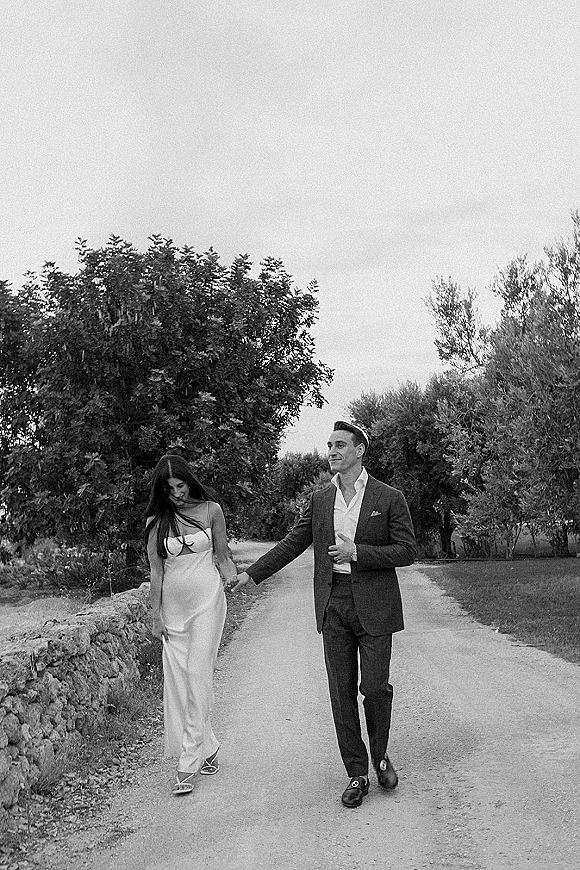 Couple portrait of bride and groom walking hand in hand, her strapless slip dress and his dark suit along a country road by a stone wall