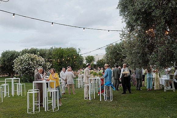 Cocktail hour setup with high top tables, string lights, floral centerpieces, and drink glasses on a lawn with trees under a cloudy sky