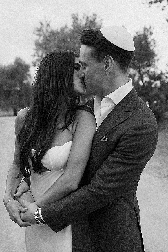 Wedding kiss portrait of bride and groom kissing, her strapless dress and his suit with kippah on a tree-lined dirt road under open sky